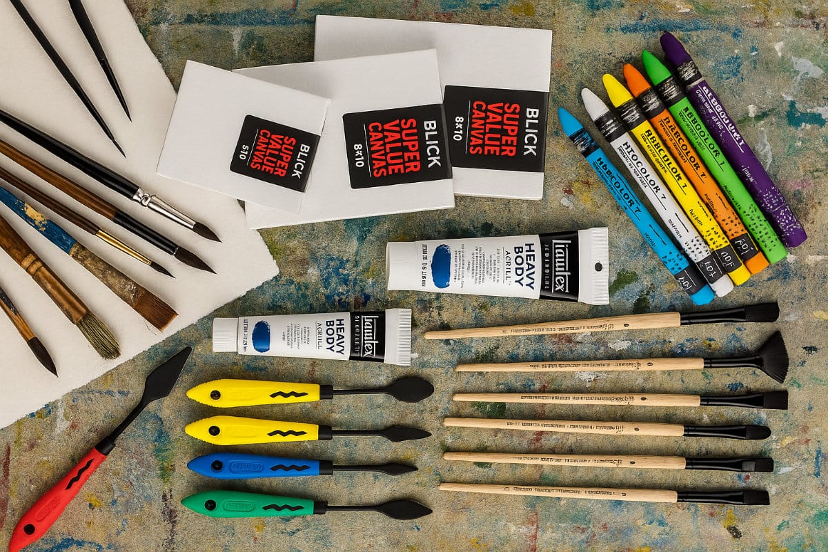 Overhead view of mixed media art supplies including pastels, paintbrushes, acrylic paints, palette knives, watercolor paper, and canvas boards on a colorful, paint-stained surface.