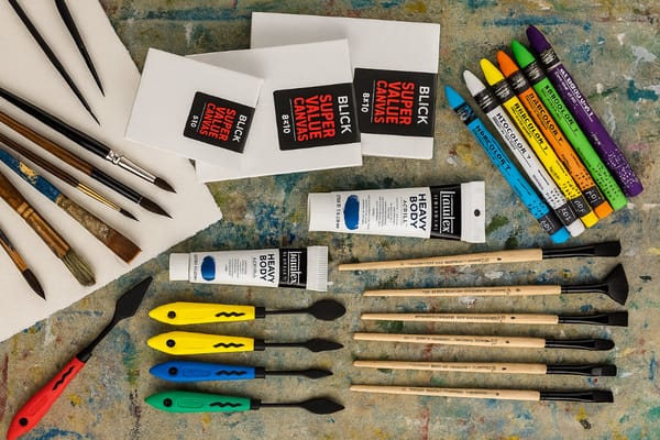 Overhead view of mixed media art supplies including pastels, paintbrushes, acrylic paints, palette knives, watercolor paper, and canvas boards on a colorful, paint-stained surface.