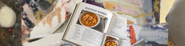 Seafood gumbo cookbook at the center of a messy mixed media artist’s desk, surrounded by collage papers.